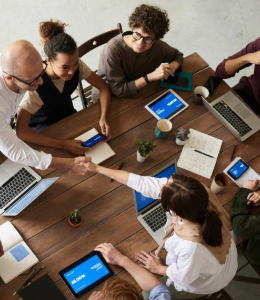 Group of diverse professionals gathered around a wooden table in a meeting, using laptops and tablets. 