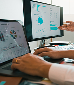 One person is typing on a laptop while another is gesturing toward a monitor displaying charts and graphs.