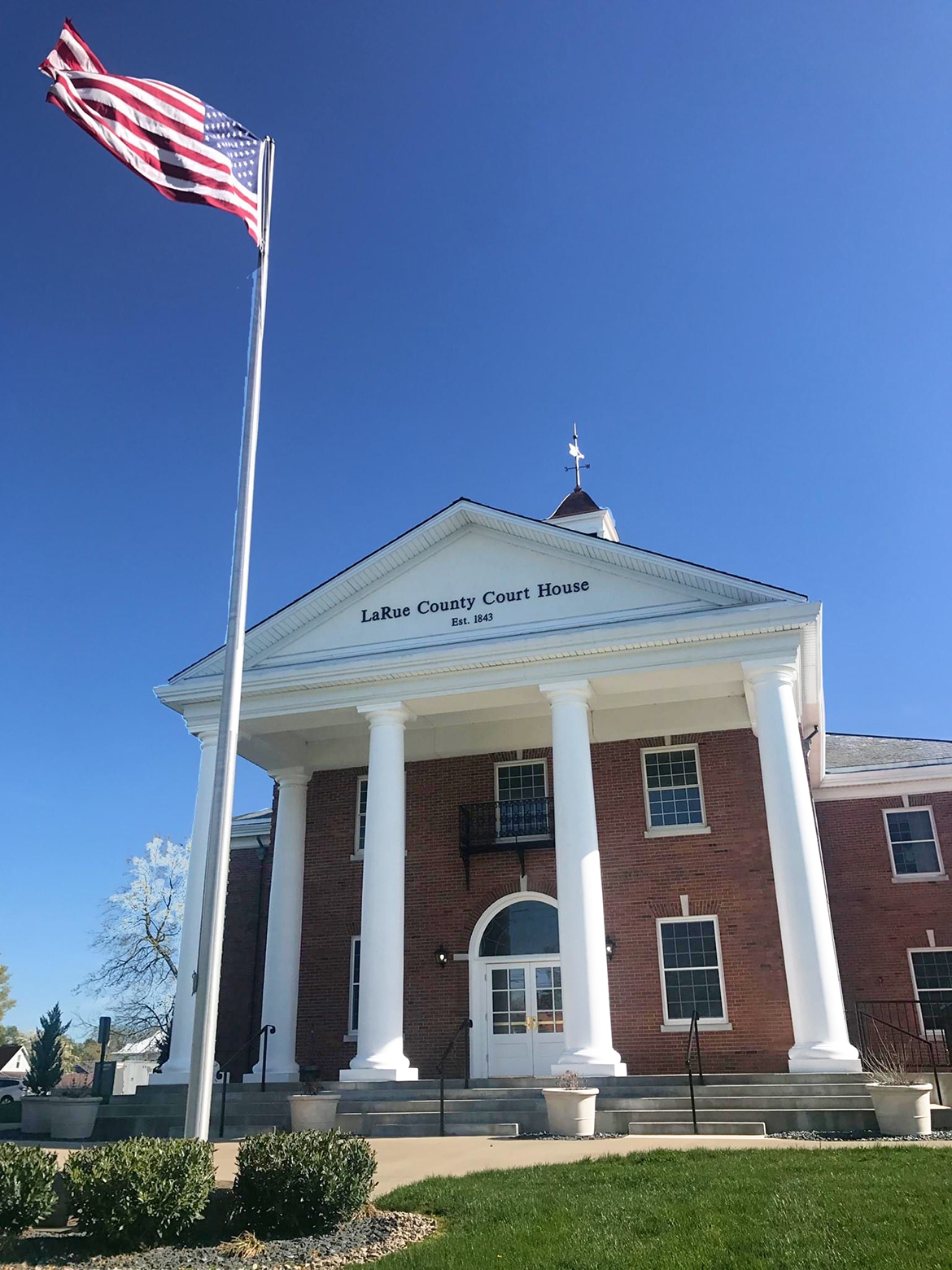 LaRue County Courthouse Annex, eaturing a grand entrance, tall columns, and a symmetrical facade. U.S.A flag on pole in front. 